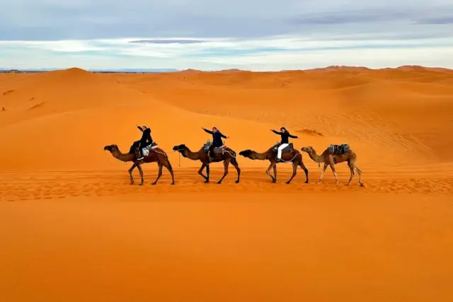 Viajeros en fila sobre dromedarios atravesando las dunas de Erg Chebbi, Merzouga, con el sol bajo